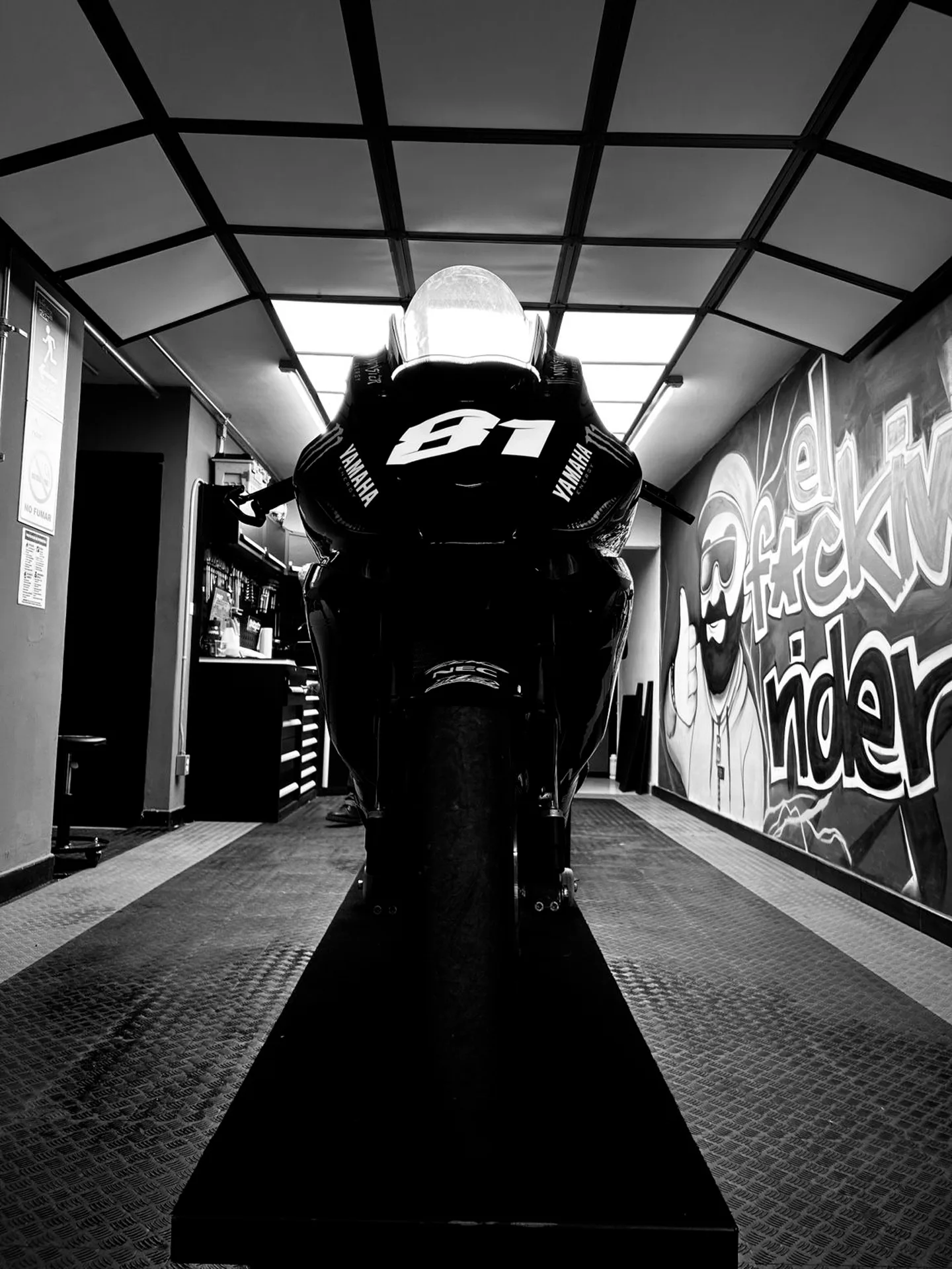 Vista frontal en blanco y negro de una motocicleta deportiva bajo el techo geométrico y junto al mural de El FKing Rider